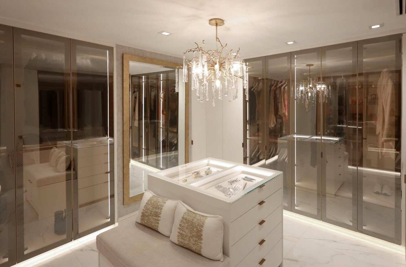 Sleek and streamlined, this gorgeous walk-in closet accented by LED strip lighting has the perfect balance of hanging, shelving and drawer space. Located in Miami Beach this client requested a modern take on the Miami Closet. Adding bronze reflective glass doors to the upgraded white finish with matte gold accessories really brought this room to life. The island with a glass top countertop for jewelry display was just the cherry on top. Photo Creds: Giang Haus